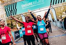 Happy female runners crossing the finish line at the Endure Relays event, celebrating their achievement with arms raised, in a vibrant outdoor setting.
