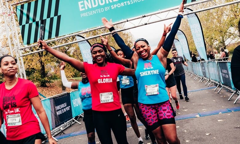 Happy female runners crossing the finish line at the Endure Relays event, celebrating their achievement with arms raised, in a vibrant outdoor setting.