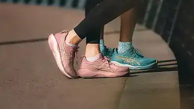 Two runners wearing pink and turquoise running shoes standing on a city sidewalk near a railing, ready for a run.