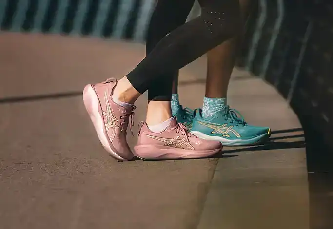 Two runners wearing pink and turquoise running shoes standing on a city sidewalk near a railing, ready for a run.