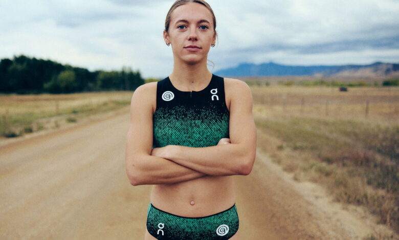 Young female runner standing confidently on a rural dirt trail with open fields and mountains in the background, wearing running gear, under cloudy skies.