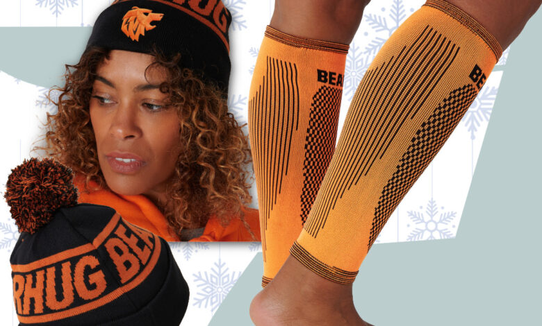 Person wearing orange knee-high socks and two knit beanies with pom-poms, set against a pale blue background with snowflake patterns.