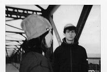 Two young runners, a girl and a boy, standing on a bridge during a running event, with the girl wearing a cap and the boy in a hoodie, in black and white.