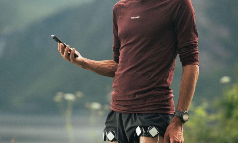 Male runner in outdoor landscape checking his smartphone, with running devices attached to his shorts, mountains in the background.