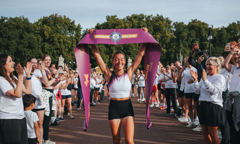 Female runner celebrating victory at a marathon finish line with a large purple ribbon, surrounded by cheering supporters on a sunny day.