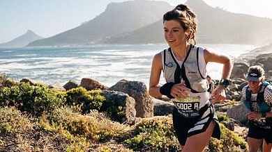 Two female trail runners competing on a rugged coastal trail with mountains and ocean in the background.