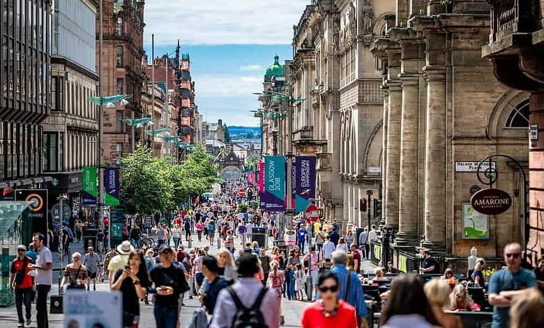 Bustling city street with pedestrians, historic buildings and storefronts featuring banners and signage, sunny day.