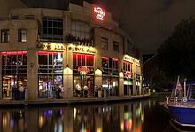 Night view of Hard Rock Cafe Amsterdam illuminated with neon lights, reflecting on the canal water, with boats docked nearby.