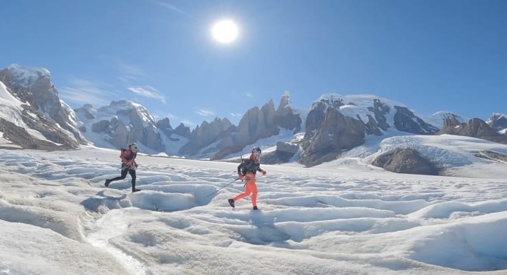 Two hikers cross a sunlit snow-covered alpine glacier with rugged peaks in the background under a clear blue sky.