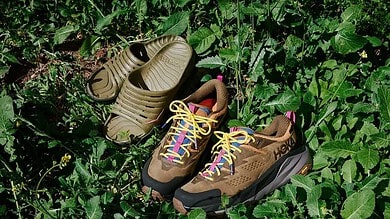 Pair of hiking trail shoes and sandals placed on green leafy ground, highlighting outdoor footwear for runners.