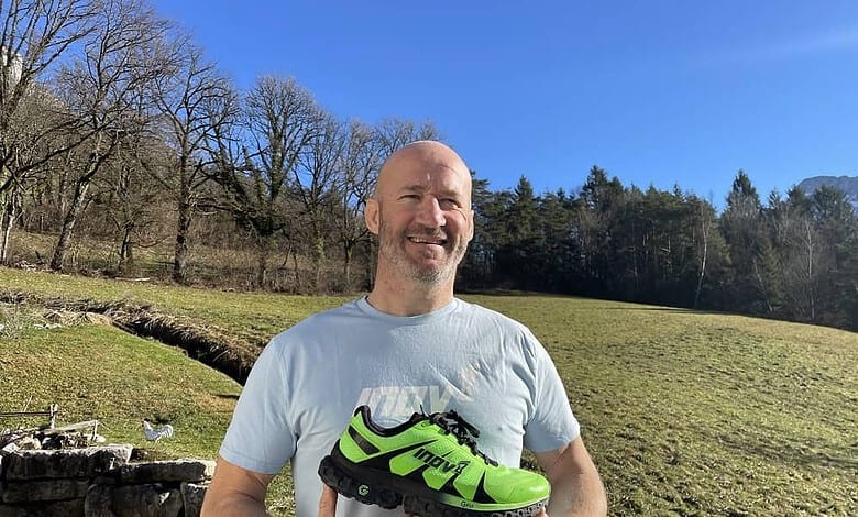 Smiling man holding bright neon green trail running shoe in an open grassy field with trees and clear blue sky, showcasing running footwear from Running Insights.