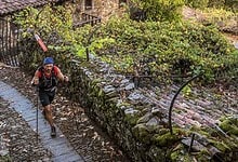 Male trail runner navigating a muddy path through a lush, green forested area with stone walls and rustic surroundings.