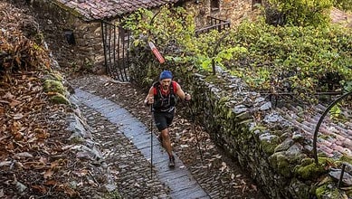 Male trail runner navigating a muddy path through a lush, green forested area with stone walls and rustic surroundings.