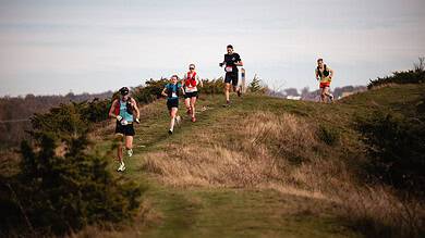 Group of runners participating in trail running on a scenic outdoor path with grassy hills and bushes under a cloudy sky.