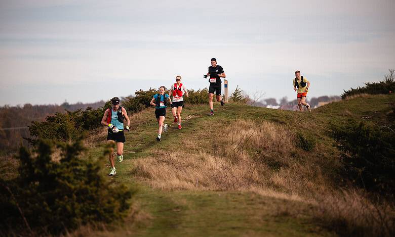 Group of runners participating in trail running on a scenic outdoor path with grassy hills and bushes under a cloudy sky.