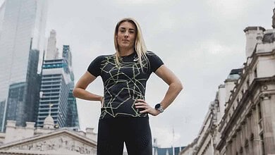 Athletic woman standing confidently in city centre, wearing a black geometric-patterned top, black leggings, and green running shoes, with hands on hips in an urban setting.