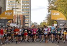 Large group of runners at the start line of a marathon in an urban setting with tall buildings and autumn trees.