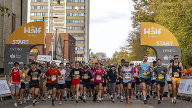 Large group of runners at the start line of a marathon in an urban setting with tall buildings and autumn trees.
