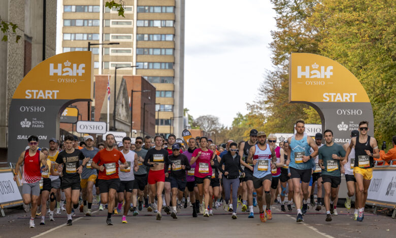 Large group of runners at the start line of a marathon in an urban setting with tall buildings and autumn trees.