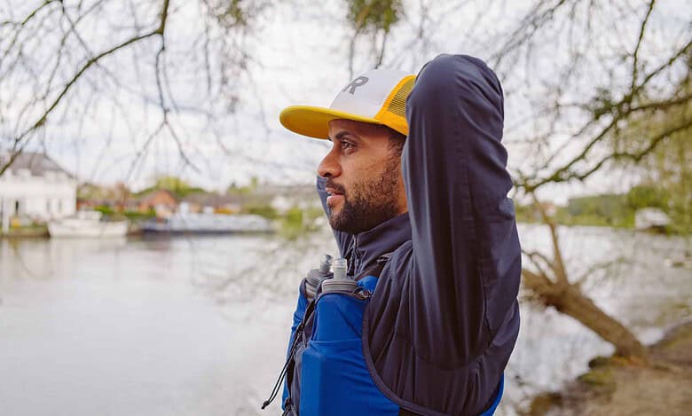 Athlete in a blue hydration vest stretching near a lake, wearing a yellow and white cap.