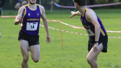 Two male runners competing in a cross country race on a grassy field, wearing purple and yellow uniforms, with trees in the background.