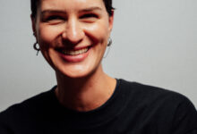 Smiling female runner with short dark hair wearing a black running shirt, showcasing confidence and fitness, against a plain grey background.