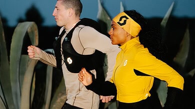 Two runners training outdoors at dusk, wearing running gear and backpacks, with large agave plants in the background, promoting running and fitness.