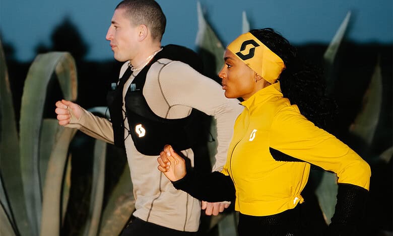 Two runners training outdoors at dusk, wearing running gear and backpacks, with large agave plants in the background, promoting running and fitness.