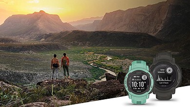 Two hikers stand on a rocky hillside overlooking a valley at sunset with a scenic mountain range in the distance and a teal watch in the foreground.