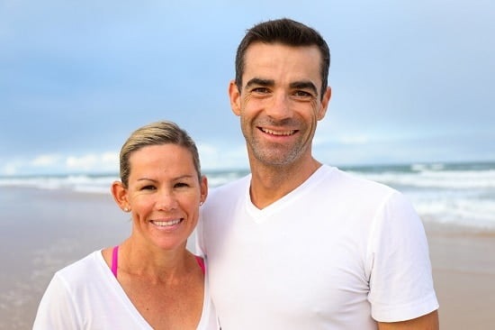 Smiling couple in white tops on a beach, waves in the background, sunlight creating a bright, uplifting scene.