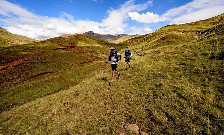 Two trail runners navigating a grassy hillside trail under a bright blue sky with scattered white clouds.