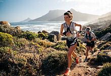 Female trail runner in a race along a rugged coastal trail with mountains and ocean in the background, wearing a race bib and athletic gear.