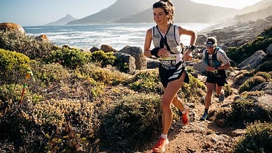 Female trail runner in a race along a rugged coastal trail with mountains and ocean in the background, wearing a race bib and athletic gear.