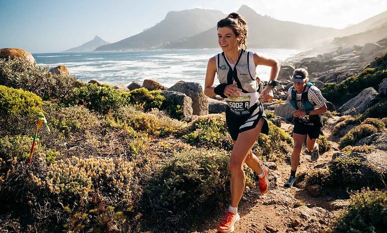 Female trail runner in a race along a rugged coastal trail with mountains and ocean in the background, wearing a race bib and athletic gear.