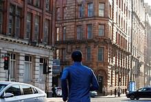 Male runner in blue jacket and black shorts running through city streets with historic buildings in the background.