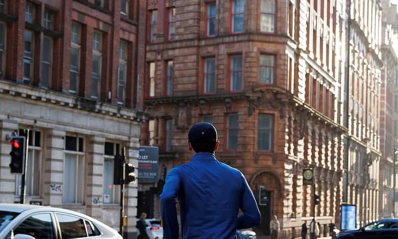 Male runner in blue jacket and black shorts running through city streets with historic buildings in the background.