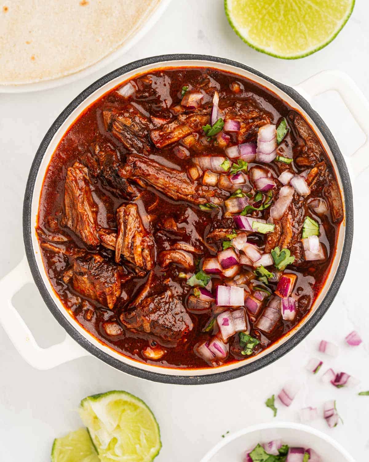 Shredded beef birria in a pot with sauce topped with diced onions and chopped cilantro.