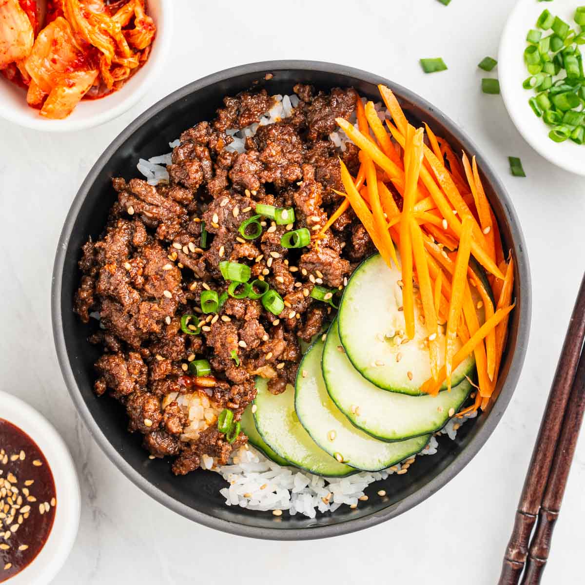 Korean beef over bowl of rice with shredded carrots and sliced cucumbers.