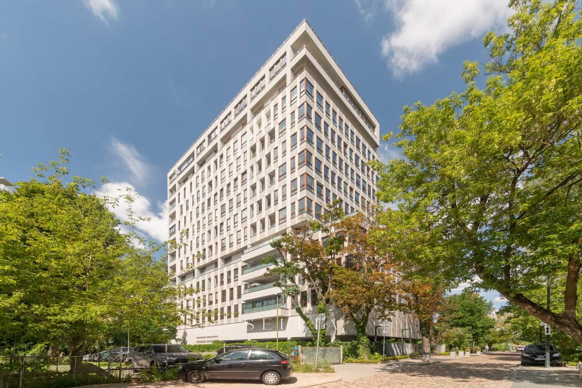 Wysoki nowoczesny apartamentowiec na Żoliborzu z dużymi oknami i zielonym otoczeniem.