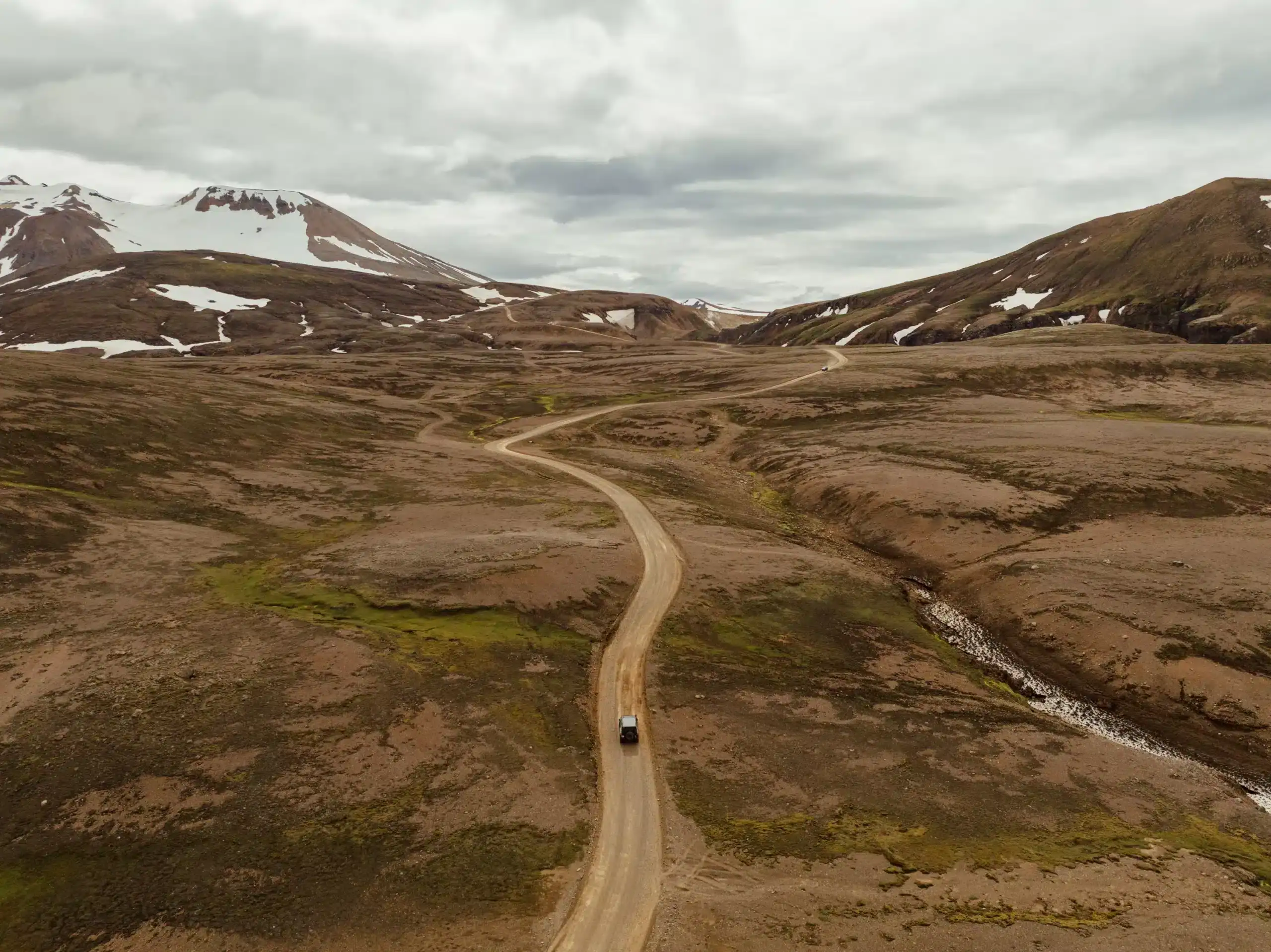A landscape of Kerlingafjoll mountains partly covered with snow and a road on the ground where two cars are transferring on the Kerlingafjöll Highland transfer.