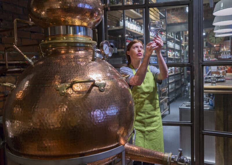 Woman in green apron inspecting copper still in distillery. at St Christopher's Edinburgh Old Town hostel