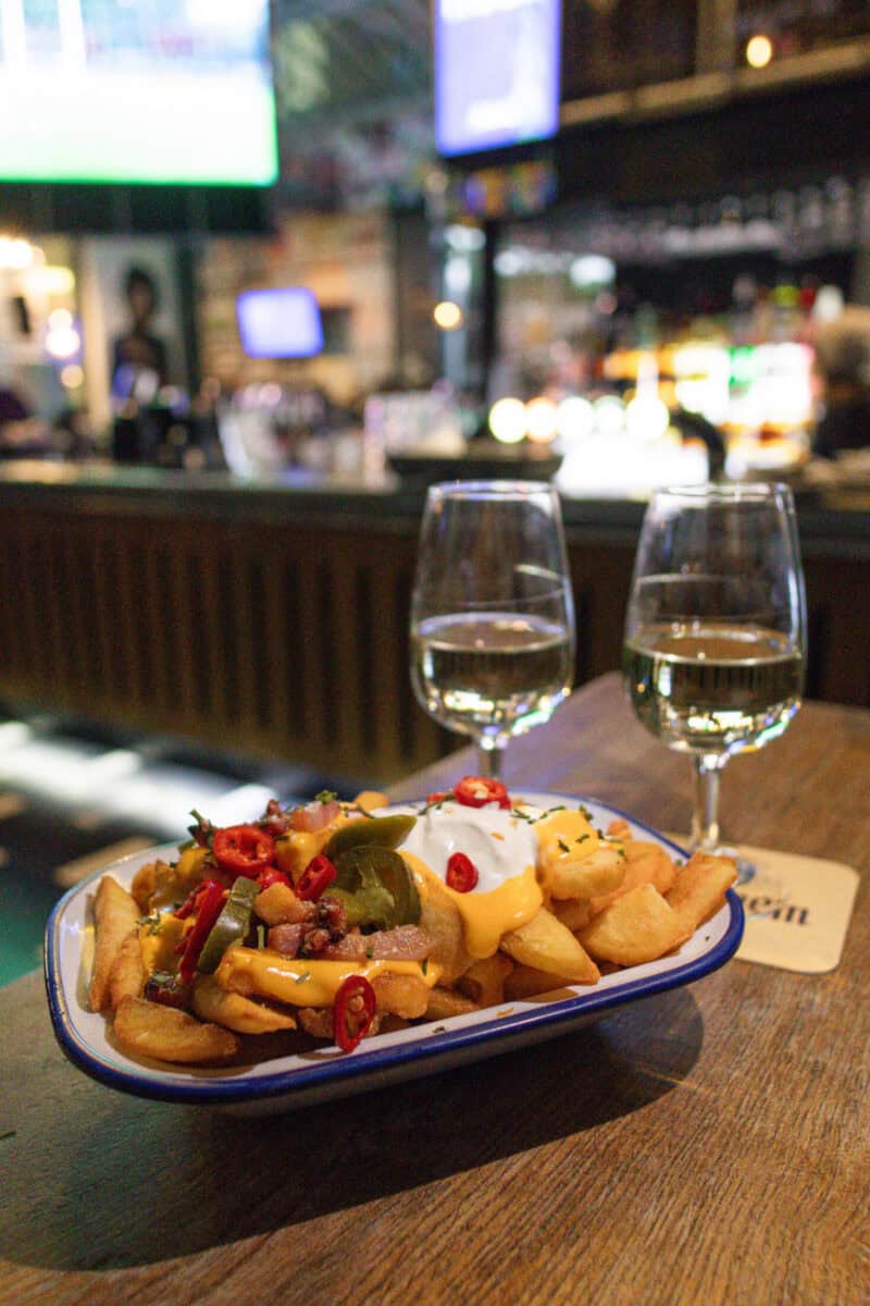 A plate of loaded fries with toppings and two glasses of white wine in a social hostel bar.