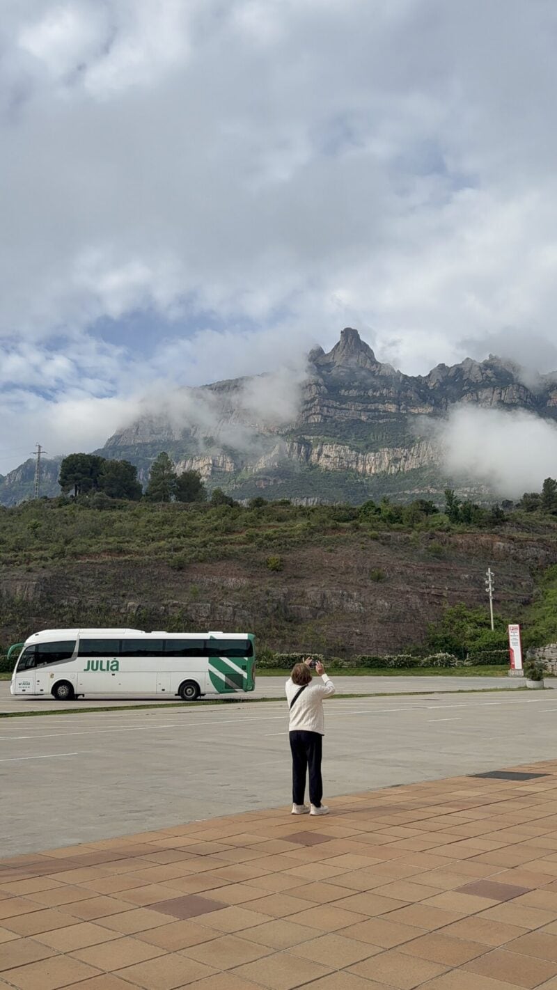 A traveller taking a photo of a scenic mountain backdrop with a bus in a large car park.