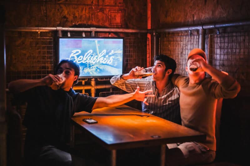 A lively social hostel bar with three young people enjoying drinks and fun.