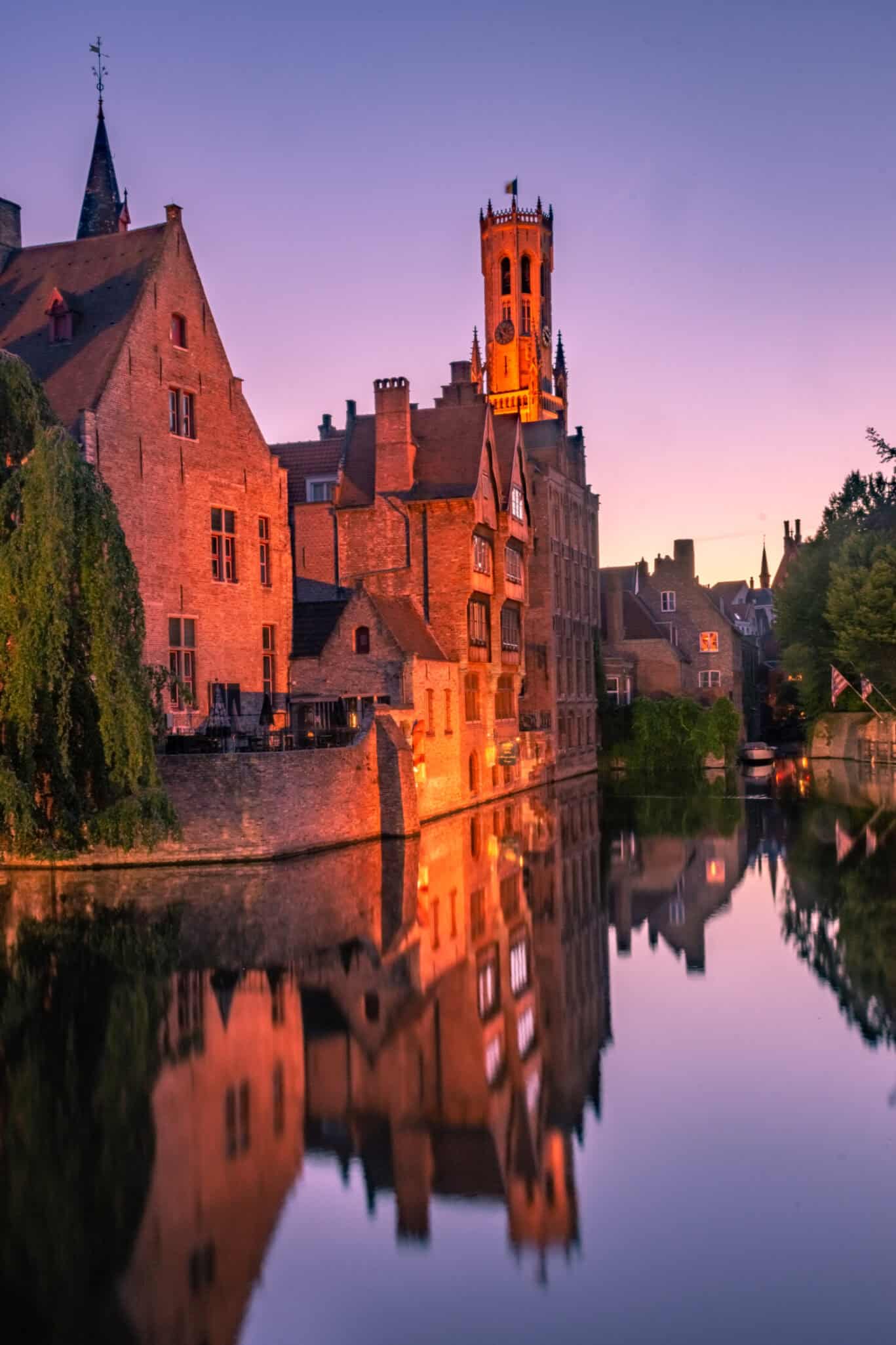 Historic town buildings and a church tower reflected in a river at sunset.