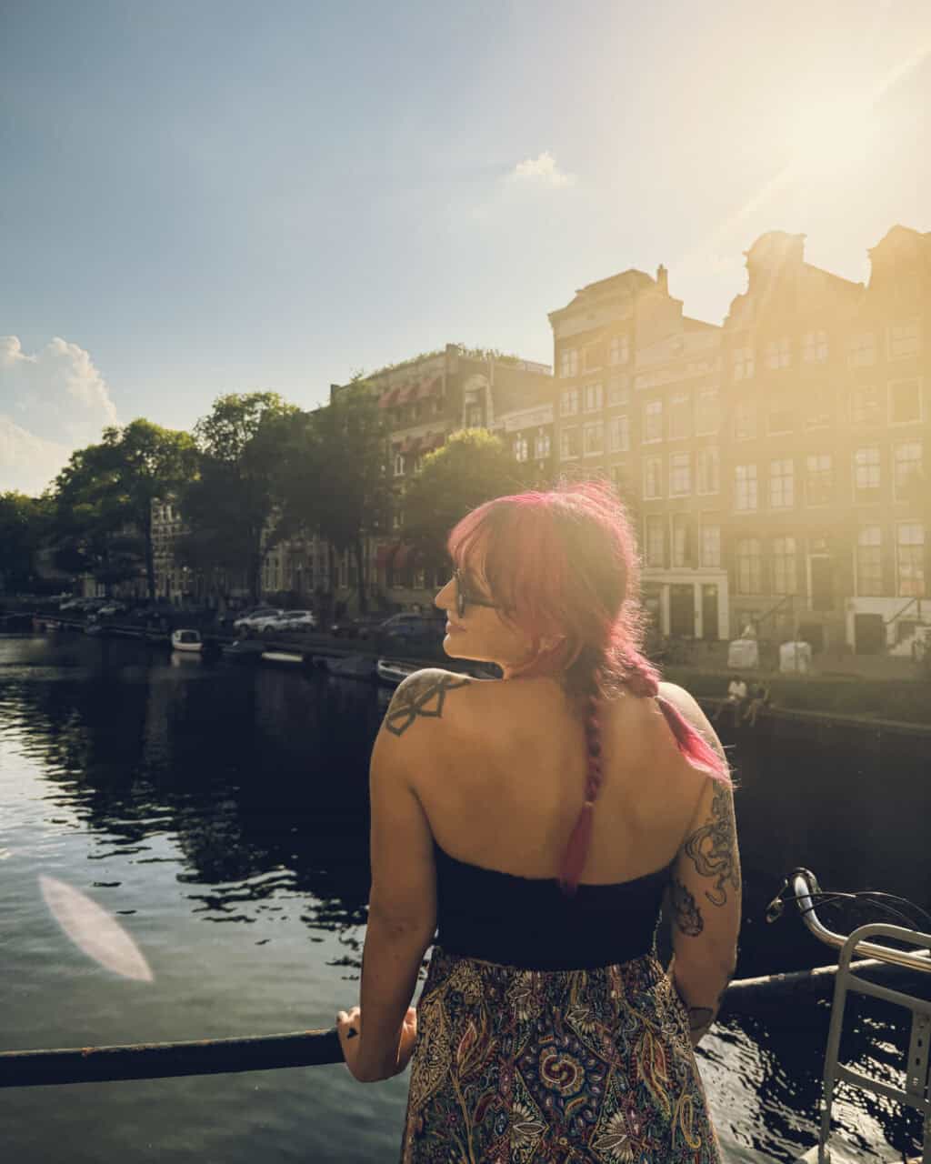A woman with pink hair and tattoos looking over a canal with historic buildings.