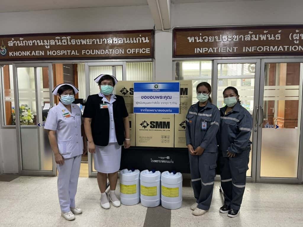 ผู้หญิงและผู้ชายในเครื่องแบบโรงพยาบาลและชุด PPE ยืนหน้าประตูสำนักงานมูลนิธิโรงพยาบาลขอนแก่น พร้อมกล่องบรรจุอุปกรณ์ทางการแพทย์และถังเก็บของเหลวในโรงพยาบาล ข้อมูลภาพแสดงถึงความร่วมมือด้านสุขภาพและการสนั.