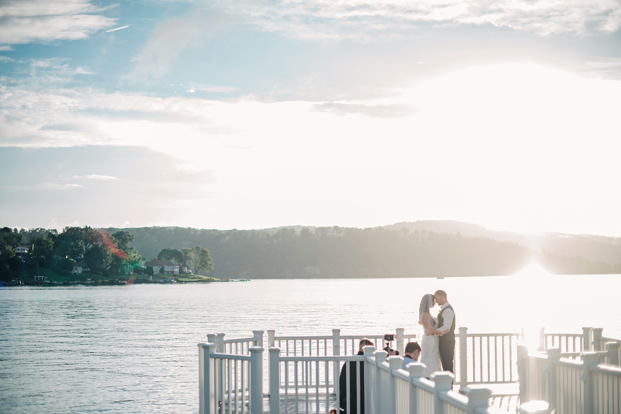Bride and groom share a kiss on a lakeside pier during a glowing sunset portrait.