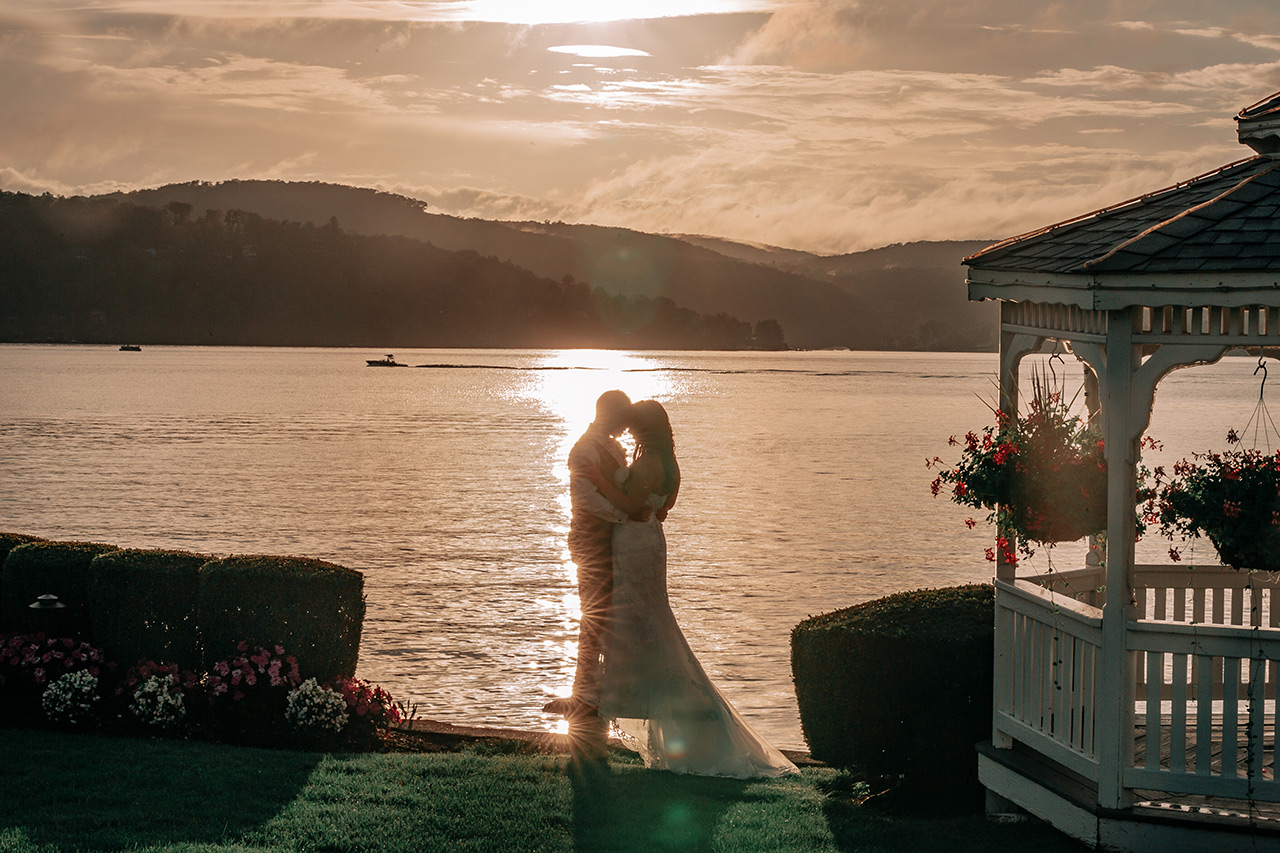 Romantic lakeside wedding portrait at sunset with couple embracing beside a gazebo.