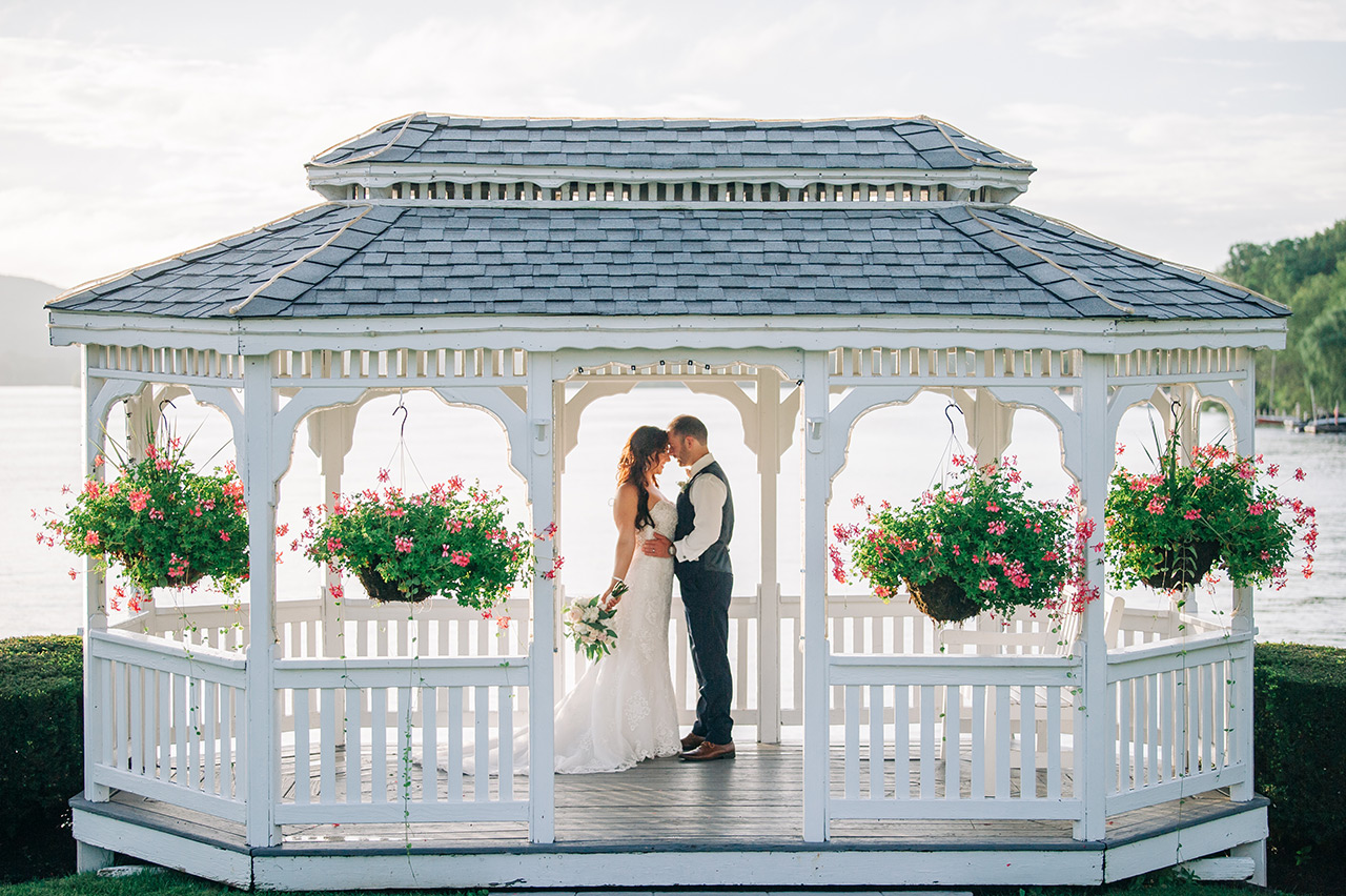 Bride and groom share a romantic moment beneath a lakeside gazebo adorned with flowers.